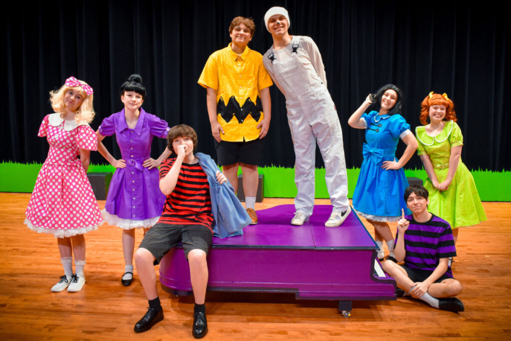 The cast of You're a Good Man, Charlie Brown gathers around Schroeder’s piano.